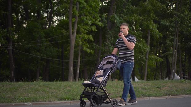 A young father carries a stroller with his son and drinks coffee in the park. A man walks with a baby in a stroller in the park. 4k