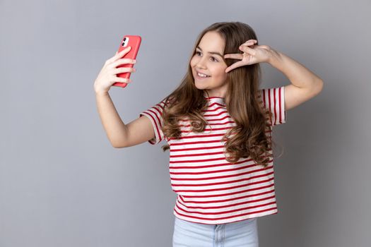 Portrait of little girl wearing striped T-shirt makes peace sign or V gesture at camera of cell phone, poses for making selfie or video call. Indoor studio shot isolated on gray background.