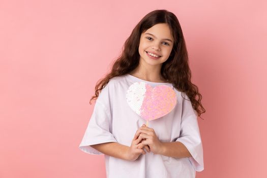 Portrait of happy satisfied little girl wearing white T-shirt holding paper pink heart on stick and smiling to camera, sharing love. Indoor studio shot isolated on pink background.