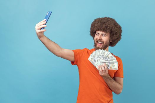 Portrait of positive optimistic man with Afro hairstyle wearing orange T-shirt having video call or livestream, showing dollar banknotes to followers. Indoor studio shot isolated on blue background.
