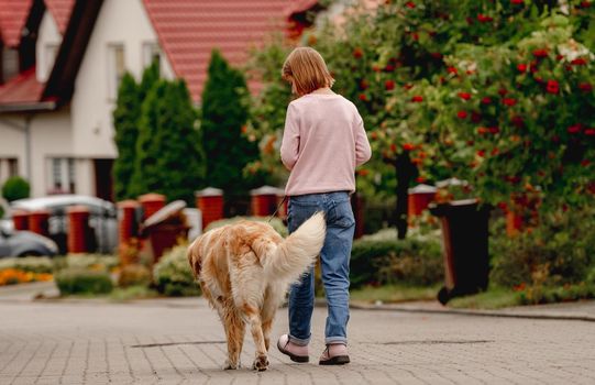 Preteen girl with golden retriever dog walking outdoors together. Back view. Pretty kid child with purebred pet doggy at street