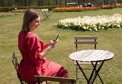 A young beautiful girl in a red dress in a spring park plays with her hair, flirts and takes a selfie on a mobile phone, tulips bloom and people walk in the park, pamper themselves.High quality photo