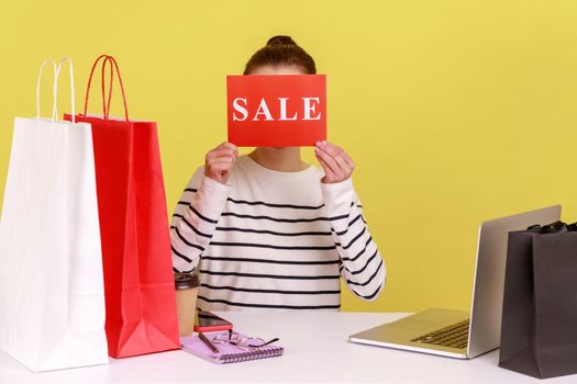 Unknown anonymous woman hiding her face behind card with sale inscription, sitting at workplace among shopping bags, huge discounts. Indoor studio studio shot isolated on yellow background.