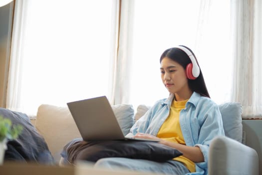 Young asian woman studying, online learing, and listening to music with headset while sitting on couch at home. Listening to podcast, audio book, watching videos using laptop. 
