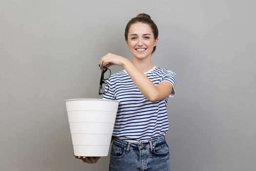 Portrait of smiling positive pretty woman wearing striped T-shirt throwing out eyeglasses to white bin, vision treatment, laser correction. Indoor studio shot isolated on gray background.