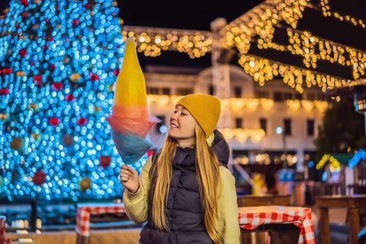 Young woman holding a cotton candy and smiling at a Christmas fair wearing a yellow wool cap.