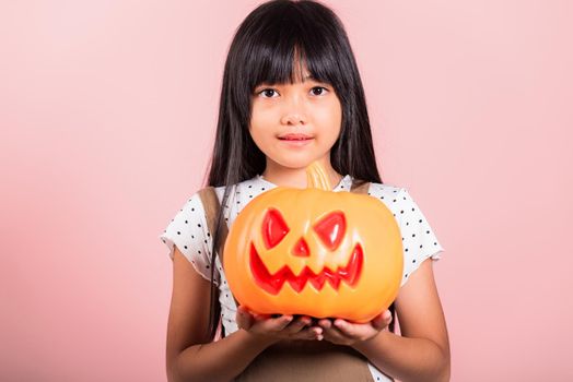 Asian little kid 10 years old holding carved Halloween pumpkin at studio shot isolated on pink background, Portrait of happy child girl smiling show Jack O pumpkin, Concept for Halloween holiday