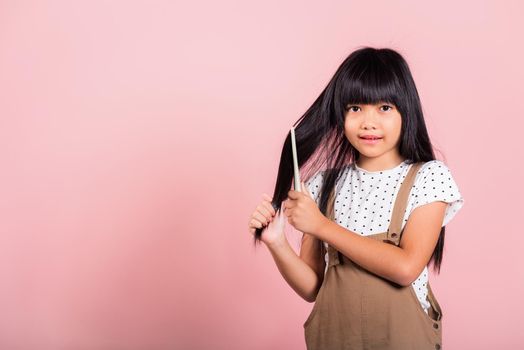 Asian little kid 10 years old hold comb brushing her unruly she touching her long black hair at studio shot isolated on pink background, Happy child girl with a hairbrush, Hair care concept