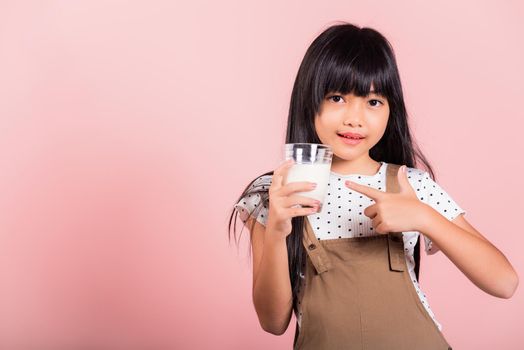 Asian little kid 10 years old smile hold milk glass drink white milk and pointing finger at studio shot isolated on pink background, Happy child girl daily life health care Medicine food