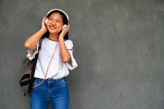 Smiling Asian female teenager listening to music with headphones on dark concrete wall background. Copy space. Lifestyle concept.