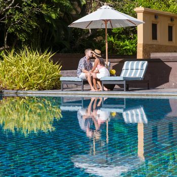 a couple of men and women relaxing by a swimming pool on a beach bed with reflections in the pool. couple on vacation relaxing on a chair by the pool