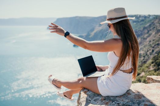 Successful business woman in yellow hat working on laptop by the sea. Pretty lady typing on computer at summer day outdoors. Freelance, travel and holidays concept.