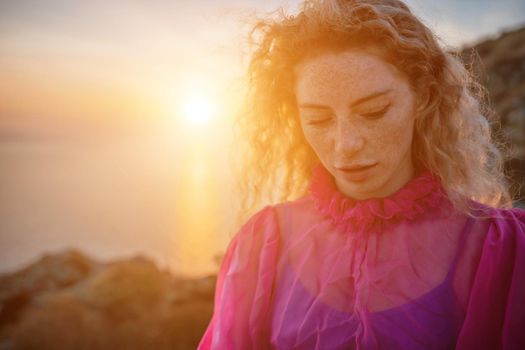 Side view a Young beautiful sensual woman in a red long dress posing on a volcanic rock high above the sea during sunset. Girl on the nature on overcast sky background. Fashion photo