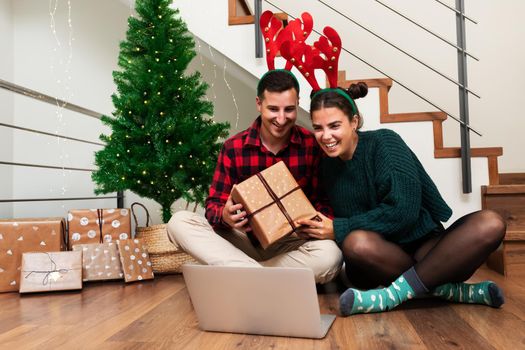 Young couple wearing felt reindeer antlers on a video call showing some Christmas presents to family. Holiday concept.