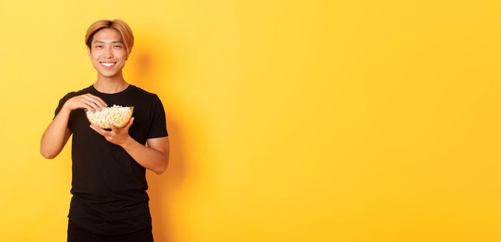 Pleased handsome korean guy smiling happy as enjoying watching movie or tv series, eating popcorn, standing yellow background.