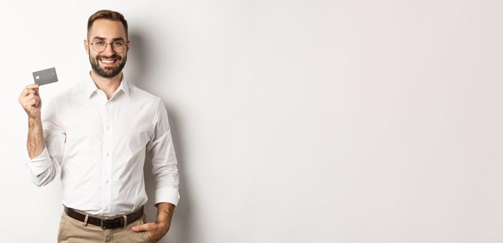 Handsome businessman showing his credit card, looking satisfied, standing over white background.