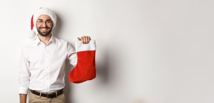 Merry christmas, holidays concept. Happy adult man receive gifts in xmas sock, looking excited, wearing santa hat, white background.