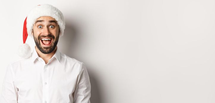 Close-up of happy bearded guy in santa hat, looking surprised, celebrating christmas, standing over white background.