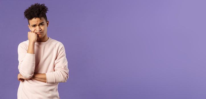 Portrait of complicated, troubled young hispanic man facing tough problem, lean on fist grimacing and pouting, solving troublesome situation, thinking, feeling uneasy, purple background.