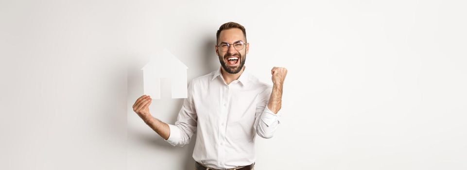 Real estate. Satisfied man rejoicing of founding perfect home apartment, holding paper house model, standing over white background.