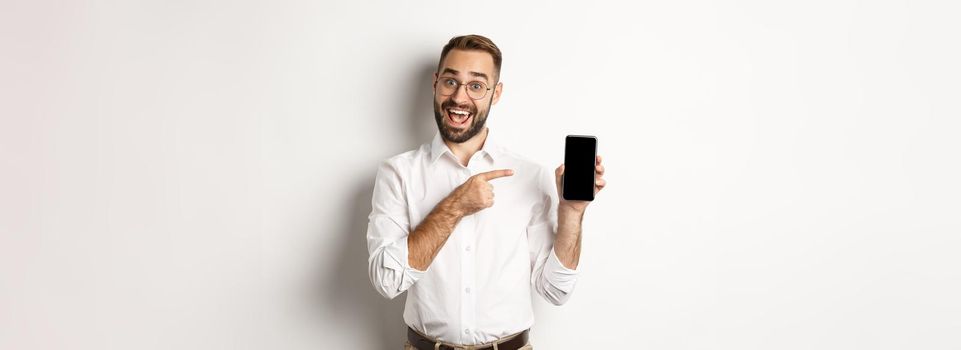 Excited handsome guy showing mobile phone, pointing finger at screen and smiling, standing against white background.