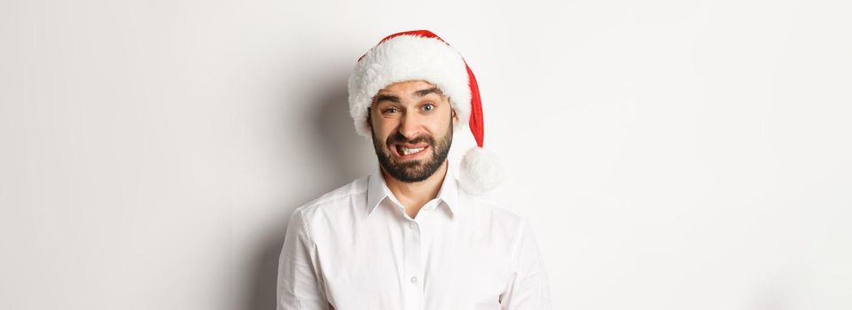 Close-up of awkward guy in santa hat apologizing, feeling uncomfortable, standing over white background. Christmas concept.