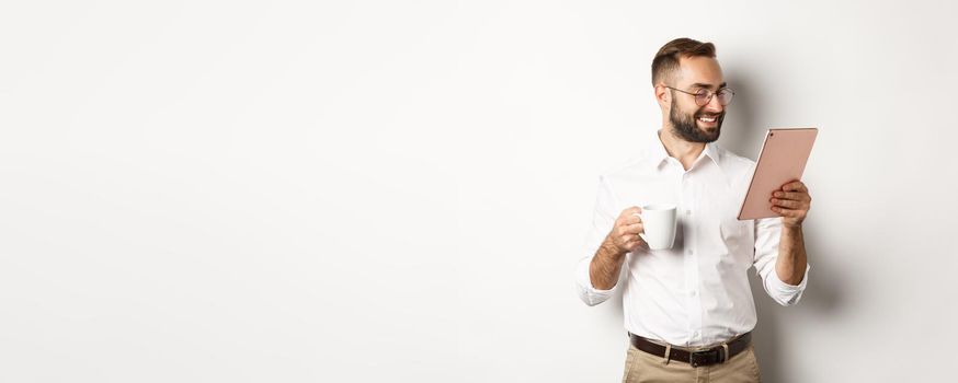 Handsome businessman drinking coffee and reading on digital tablet, smiling pleased, standing over white background.