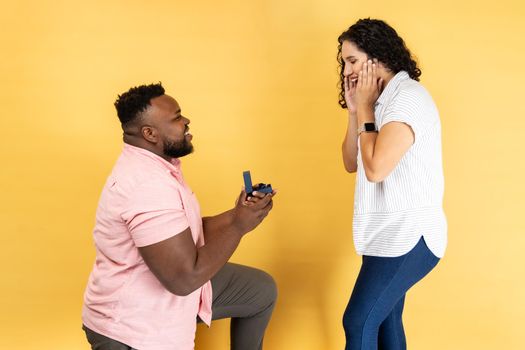 Portrait of positive young couple in casual clothing standing together, man stands on knee and making proposal to her girlfriend. Indoor studio shot isolated on yellow background.
