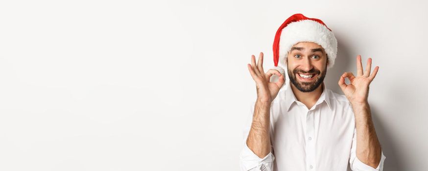 Party, winter holidays and celebration concept. Joyful man enjoying christmas and showing okay sign, smiling satisfied, wearing santa hat, white background.