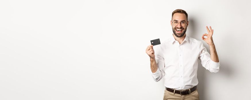 Handsome man showing his credit card and okay sign, recommending bank, standing over white background. Copy space