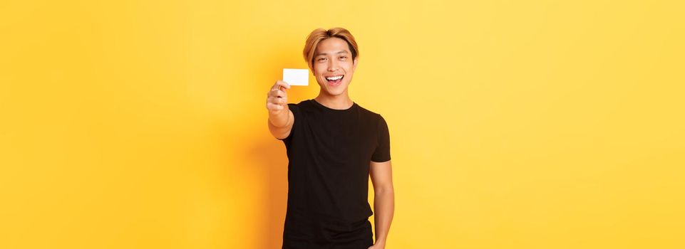 Cheerful handsome asian guy smiling pleased and showing credit card, standing yellow background.