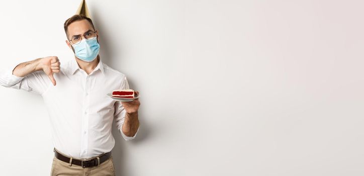 Covid-19, social distancing and celebration. Disappointed birthday guy wearing face mask, holding bday cake with wish candle, making thumb down to express dislike, white background.