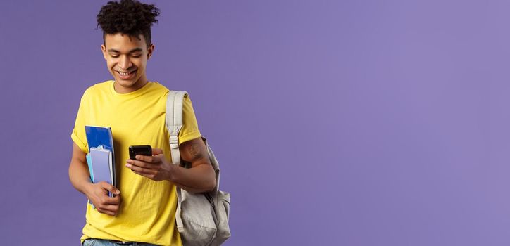 Back to school, university concept. Portrait of pleased good-looking male student texting friend, looking at mobile phone on his way to classes in campus, hold notebooks and backpack.