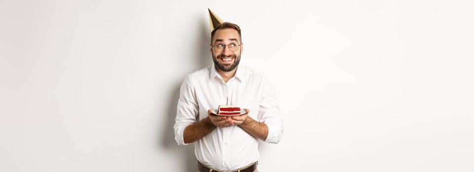 Holidays and celebration. Happy man having birthday party, making wish on b-day cake and smiling, standing against white background.