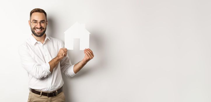 Real estate. Handsome man showing house model and smiling, broker showing apartments, standing over white background.