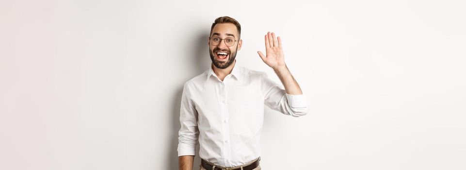 Friendly smiling man in glasses saying hello, waving hand in greeting, standing over white background.