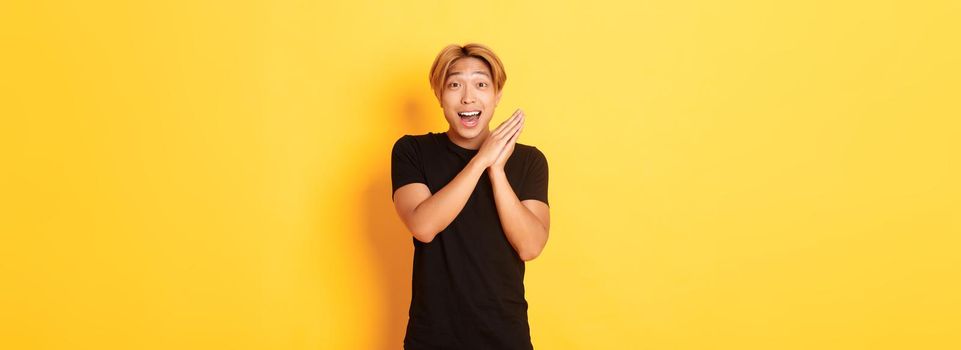 Portrait of happy and surprised asian man clap hands and smiling delighted, standing yellow background.