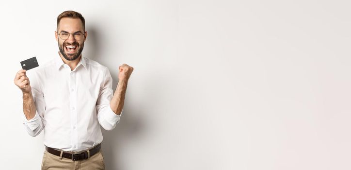 Satisfied handsome man showing credit card and rejoicing, winning something or triumphing, standing over white background.