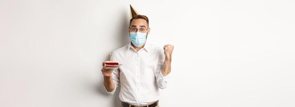 Covid-19, social distancing and celebration. Hopeful happy birthday man in face mask, holding bday cake and rejoicing, standing against white background.