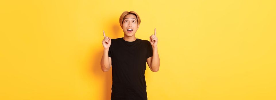 Impressed good-looking asian blond guy, wearing black t-shirt, looking and pointing fingers up excited, yellow background.