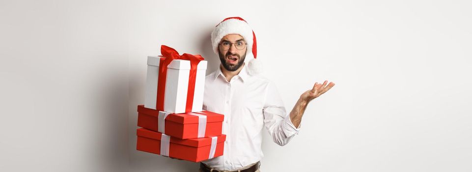 Merry christmas, holidays concept. Man looking confused while holding xmas gifts, shrugging puzzled, standing in santa hat against white background.