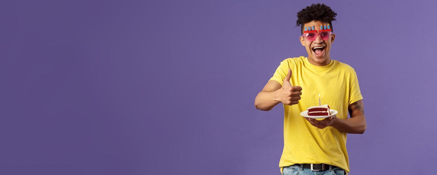 Celebration, party and holidays concept. Portrait of upbeat, cheerful young happy man celebrating birthday, wear funny sunglasses and hold b-day cake with lit candle, show thumb-up, recommend.