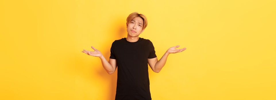 Portrait of clueless asian blond guy, wearing black clothes, shrugging and looking puzzled, standing yellow background.