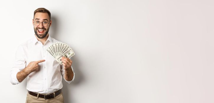 Excited businessman pointing at money, showing dollars and smiling, standing over white background.