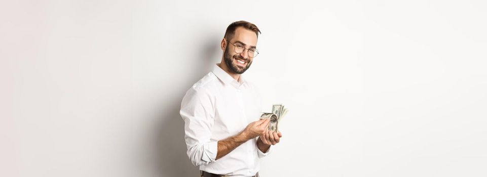 Successful business man counting money and smiling, standing against white background and looking satisfied.