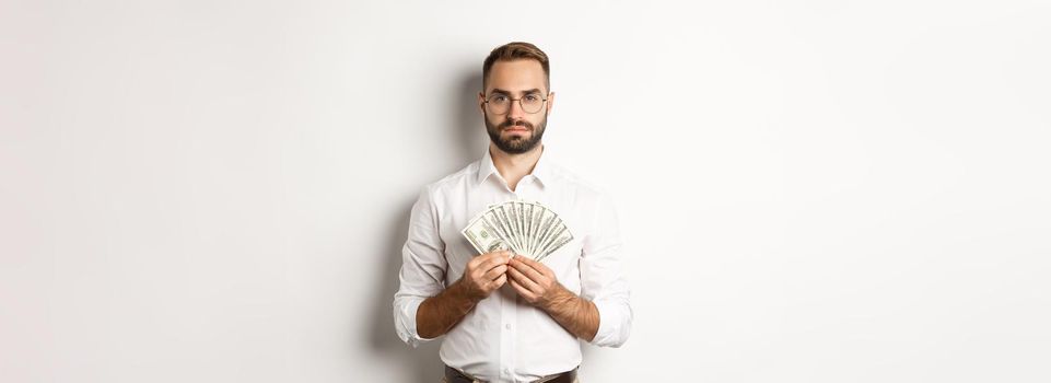 Serious businessman holding money, showing dollars, standing over white background.