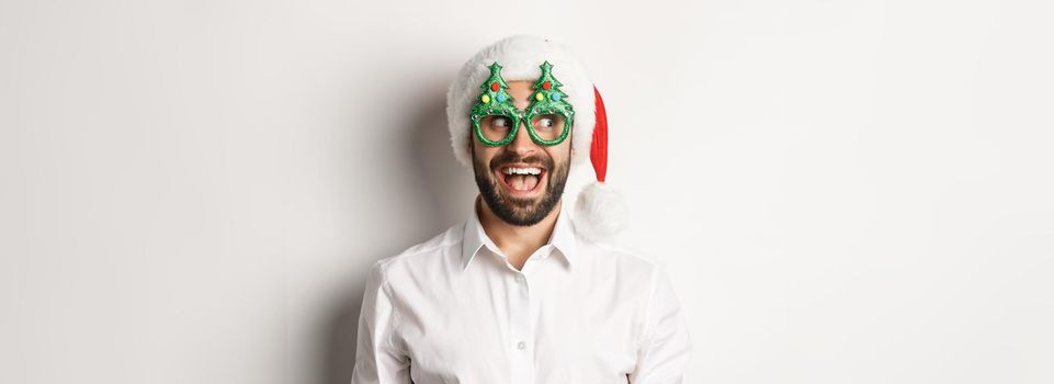 Close-up of funny man looking left with surprised face, wearing christmas party glasses and santa hat, celebrating New Year, white background.