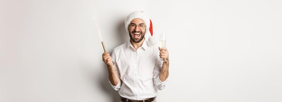 Winter holidays and celebration. Handsome bearded man having New Year party, holding firework sparkle and champagne, wearing santa hat, white background.