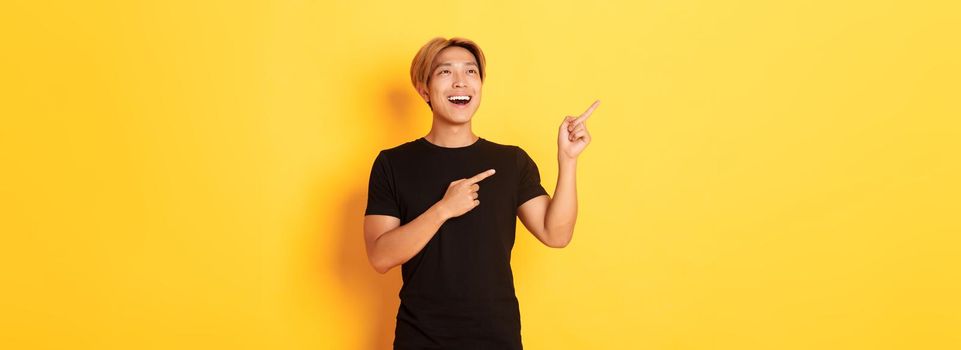 Portrait of pleased handsome asian guy in black t-shirt, pointing fingers and looking upper left corner with satisfied smile, yellow background.