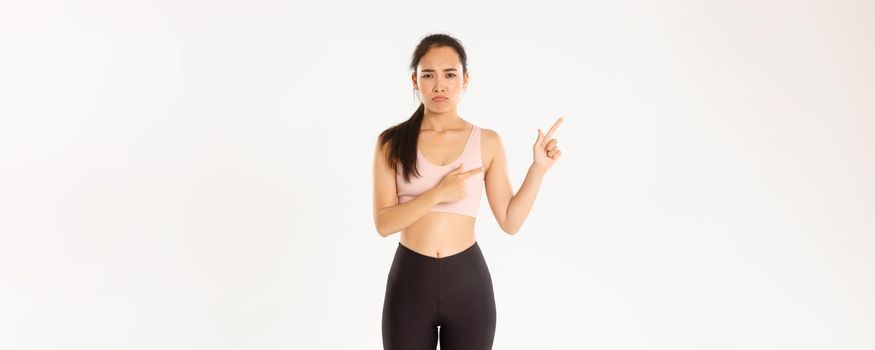 Sport, wellbeing and active lifestyle concept. Complaining sad asian girl in activewear, sulking and frowning upset as pointing upper left corner, end of sale offer, standing white background.